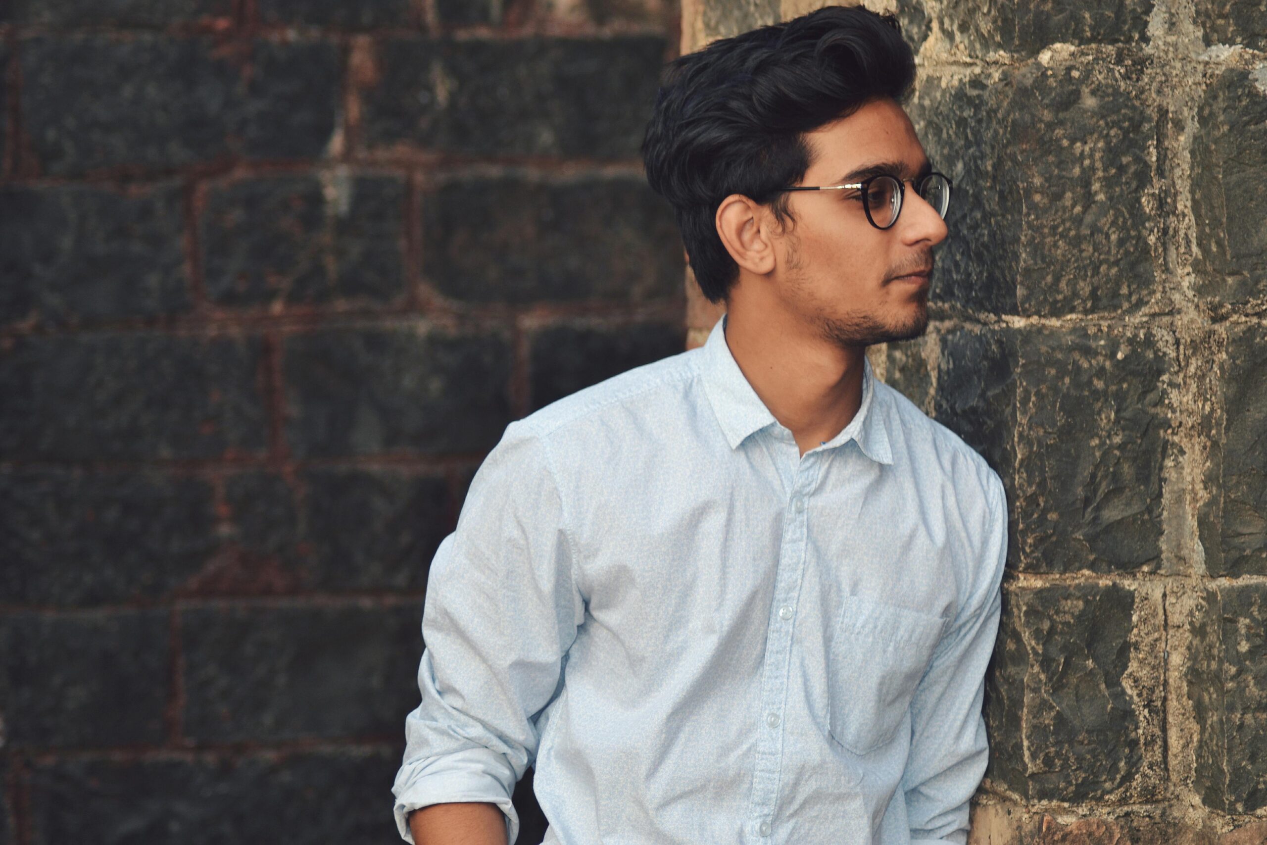 A stylish young man in glasses leans against a brick wall wearing a casual light blue shirt.